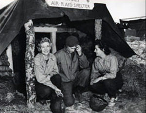 Ernie Pyle sits with two nurses in front of an air raid building.